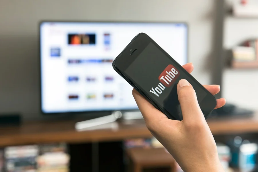 Pessoa-segurando-o-celular-aberto-em-pagina-do-YouTube-com-televisao-ao-fundo.webp